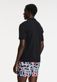 Čierne polo tričko s krátkymi rukávmi, doplnené farebnými plážovými šortkami s geometrickými vzormi v modrej, ružovej a čiernej farbe.