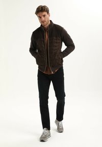Bruine ritsjack met een geborduurd voorfront, effen mouwen en hoge kraag. Draag het over een bruine blouse en gecombineerd met donkere jeans en grijze sneakers.