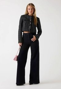 Gekürzte schwarz-weiß karierte Jacke mit langen Ärmeln, kombiniert mit hoch taillierten schwarzen Jeans mit weitem Bein, hält eine kleine pinke Handtasche.