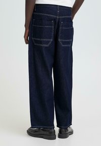 Donkerblauwe jeans met wijde pijpen en een ruwe zoom, twee achterzakken, contrasterende stiksels en voorste lusjes voor een riem. Glad denimtextuur.