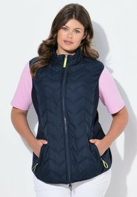 Chaleco acolchado azul marino con patrón de chevrón, cuello alto, cremallera frontal y dos bolsillos laterales. Combinado con una blusa de manga corta rosa claro.