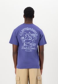 Lilla bomulls t-skjorte med et hvitt grafisk design på baksiden, som viser en tegneseriefigur med teksten "CLEPTOMANIC" og "Boule & Pastis Club."