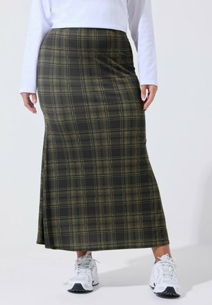 Frau, die einen langen olivgrünen karierte Rock trägt, kombiniert mit weißen Sneakers und einem weißen langärmeligen Oberteil, steht auf einem weißen Boden.