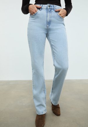 Kvinde iført lyseblå jeans med høj talje og lige ben, med hænderne i lommerne og brune spidse sko på en grå gulv.