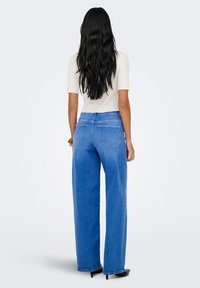 Mėlyni plačiomis kojomis džinsai vidutinio plovimo, su užpakalinėmis kišenėmis ir šiek tiek nudylančia apačia. Derinami su aptemptu trumpomis rankovėmis kremine palaidine.