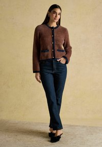 Brūns adīts kardigāns ar tumši zilām apmalēm, zelta pogām un priekšējiem kabatām, kombinēts ar tumšiem džinsiem un melniem mūliem ar loku detaļu.