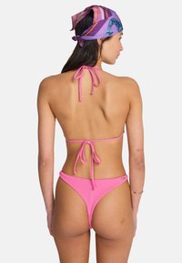 Bikini rosa a coste con dettagli annodati al collo e sulla schiena. Presenta una copertura minima e un motivo di tessuto strutturato. Sciarpa di seta in tonalità viola.