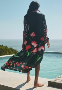 Black floral kimono with red and pink flower pattern, sheer fabric, loose fit, ankle-length, flowing sleeves, worn by a person near water.