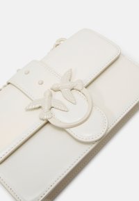 Borsa in ecopelle avorio con pattina decorata da due accenti a forma di uccello e un anello circolare. Texture liscia con dettagli di cucitura a contrasto.
