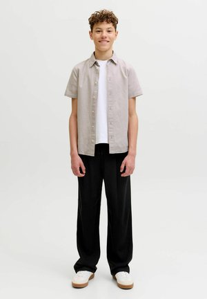 Kõrvalestadega teismeline seisab ja naeratab, seljas beež lühikeste varrukatega nööbitav särk üle valge t-särgi, mustad püksid ja valged tossud.