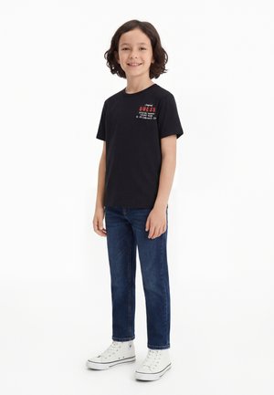 Enfant souriant aux cheveux ondulés et foncés portant un t-shirt noir GUESS, un jean bleu et des baskets blanches montantes, debout sur un fond blanc.