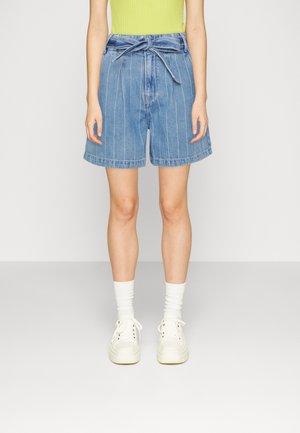 Vrouw draagt een hooggetailleerde blauwe jeansshort met fijne streepjes en een gebonden riem, witte sokken, witte sneakers en een groene mouwloze top.