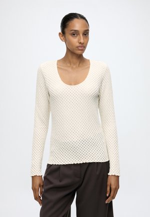 Donna con capelli scuri raccolti in uno chignon, indossa una maglia bianca lavorata a maniche lunghe e pantaloni scuri e larghi, in piedi su uno sfondo chiaro.