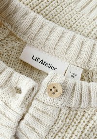 Stickad textil i krämfärg med ribbad krage och knapp. Labeln läser "Lil’Atelier," vilket indikerar storlek 1-2M. Naturlig, mjuk textur.