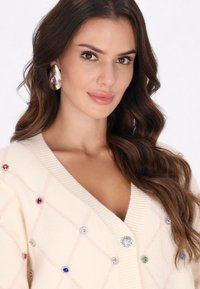Krémový cardigan s diamantovým vzorem, zdobený barevnými drahokamy a květinovým knoflíkem. Texturovaná tkanina a hluboký výstřih do V.