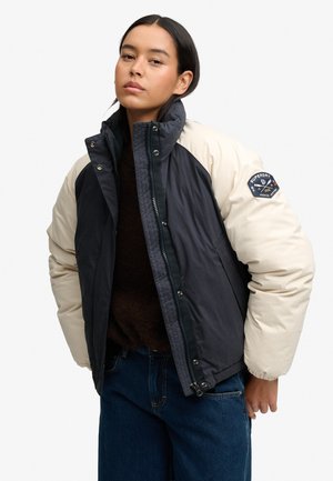 Mujer joven con una chaqueta acolchada negra y beige con un parche del logo de Superdry en la manga izquierda y vaqueros azules.