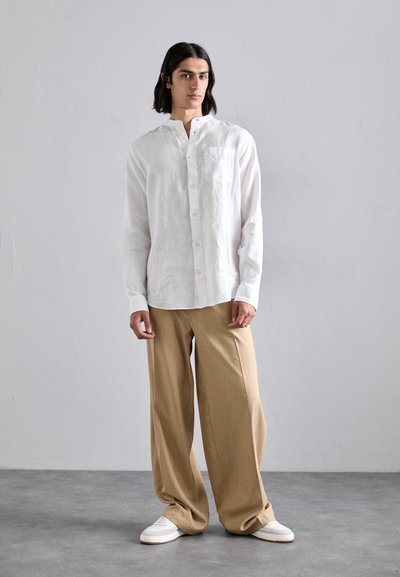 Camicia bianca in lino con colletto alla mandarino, maniche lunghe e taschino sul petto; abbinata a pantaloni larghi beige e sneakers bianche.