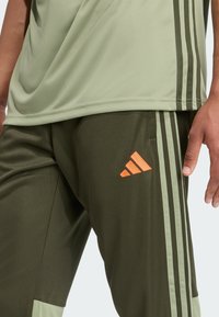Šviesiai žalia sportinė marškinėliai su laisvu siluetu ir lygiu audiniu, derinami su tamsiai žaliais kelnėmis, turinčiomis oranžinį "Adidas" logotipą ir trimis juostelėmis.