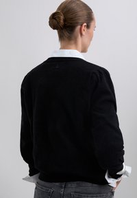 Eine Frau mit einem Dutt trägt einen schwarzen Pullover über einem weißen Hemd mit Kragen und kombiniert diesen mit dunkelgrauen Jeans. Sie steht mit dem Rücken zu einem schlichten Hintergrund.