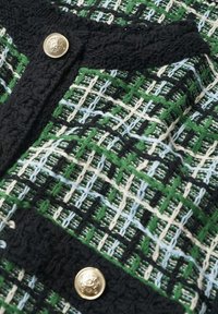 Tissu tissé vert, noir, blanc et bleu avec un bord texturé noir et deux boutons décoratifs dorés.