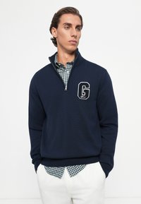 Young man wearing a navy blue quarter-zip sweater over a green and white checkered shirt, paired with white pants, standing with hands in pockets.