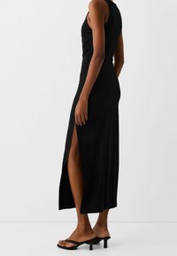 Černé bezramínkové maxi šaty s bočním rozparkem; vyrobené z hladkého materiálu s přiléhavým střihem. Malé černé sandály na podpatku doplňují vzhled.