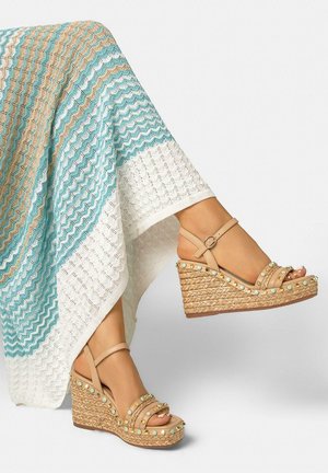 Piernas con sandalias de cuña beige con tachuelas turquesas, combinadas con una falda larga de punto con patrones de zigzag en blanco, turquesa y beige.