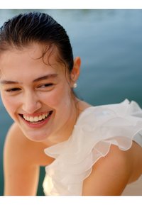 Robe blanche à volants avec tissu transparent superposé ; le mannequin a les cheveux mouillés, sourit et porte une boucle d'oreille en perle ; en arrière-plan, on voit de l'eau.