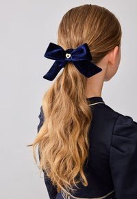 Fascetta di velluto blu navy con un accento a forma di cuore dorato, posizionata alla base di lunghi capelli castani ondulati su uno sfondo neutro.