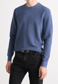 Maglione azzurro a maglia con scollo rotondo, superficie texturizzata, maniche lunghe e un piccolo logo sul petto, abbinato a jeans neri.