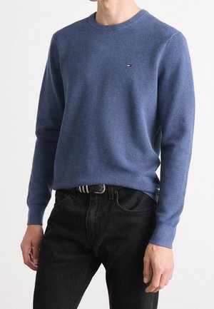 Blauer gestrickter Pullover mit runder Halslinie, strukturierter Oberfläche, langen Ärmeln und einem kleinen Logo auf der Brust, kombiniert mit schwarzen Jeans.