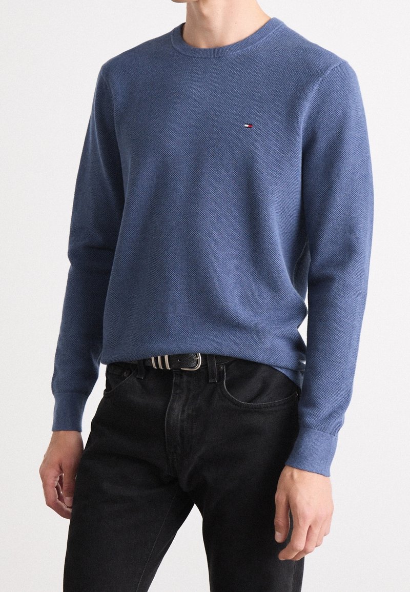 Maglione azzurro a maglia con scollo rotondo, superficie texturizzata, maniche lunghe e un piccolo logo sul petto, abbinato a jeans neri.