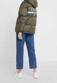 Khaki puffová bunda s kapucí, zdobená modro-šedými pruhy a logem na zádech. Spárována s modrými džíny volného střihu a bílými teniskami.