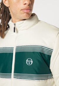 Puff jakke i creme med et grønt panel med vandrette striber. Den har en høj krave og et broderet logo på brystet. Lynlåsdetalje synlig.