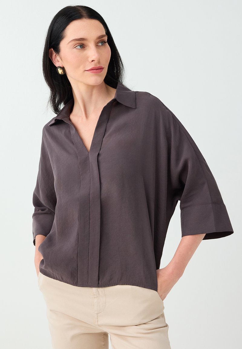 Femme aux cheveux raides foncés portant une chemise ample marron foncé à col et un pantalon beige, les mains dans les poches, regardant sur le côté.
