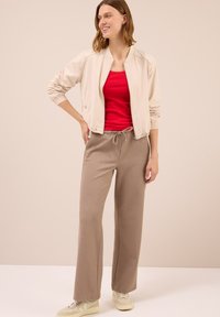Giacca bomber beige chiaro con polsini elastici, canotta rossa aderente e pantaloni della tuta marroni larghi. Le scarpe presentano una tonalità gialla chiara e un design testurizzato.
