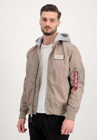 Hele beeži bomber-jakk halli kapuutsiga, lukuga sulgemine, ribakäised ja käepärane tasku, millel on punane silt. Sile tekstuur ja voolujooneline disain.