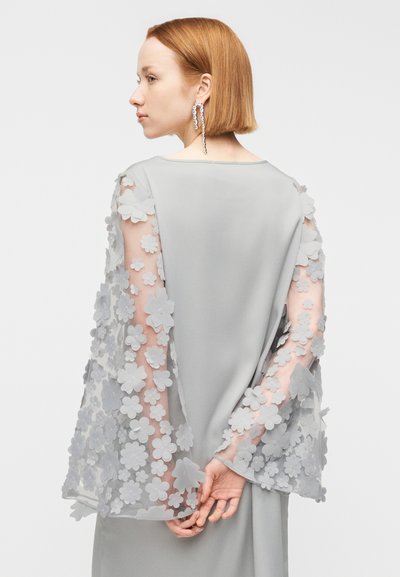 Γυναίκα με κοντά κόκκινα μαλλιά που φοράει ανοιχτό γκρι φόρεμα με διάφανα μανίκια διακοσμημένα με τρισδιάστατες ανθικές εφαρμογές, με τα χέρια σταυρωμένα πίσω από την πλάτη της.