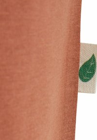 Tkanina v rjavi barvi z gladko teksturo, ki prikazuje majhen zeleni listni logotip na svetlo rjavi etiketi. Oblikovanje je preprosto in moderno.