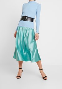 Himmelblaues, geripptes Langarmoberteil mit Rüschenakzenten, kombiniert mit einem glänzenden, türkisfarbenen Satin-Midirock und einem breiten schwarzen Gürtel. Schwarze, schmale Absatzsandalen.