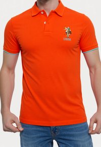 Orange polo skjorta i bomull med krage, korta ärmar och blå detaljer. Har en detaljerad broderad logotyp på bröstet.