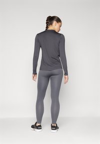Siva dolgi rokav in sivi rebrasti leggingi, oba prilegajoča. Leggingi imajo teksturiran vzorec. Model nosi črne športne čevlje.