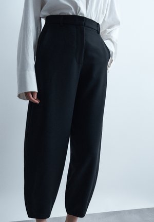 Άτομο που φοράει μαύρο παντελόνι με φαρδιά πόδια και ψηλή μέση και λευκό πουκάμισο με μακριά μανίκια και εκτεταμένες μανσέτες, στέκεται μπροστά σε απλό φωτεινό φόντο.