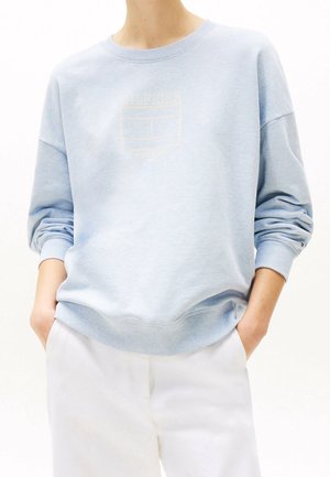 Person iført lyseblå sweatshirt med subtilt Tommy Hilfiger-logo og hvide bukser, hænder i lommerne, mod en ensfarvet baggrund.