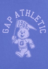 Mosolygó medve maszkot visel, amelyen "G" betű látható, egy zászlót tart, ami "1969" feliratú, a "GAP ATHLETIC" írás pedig ívben helyezkedik el a kék háttér előtt.