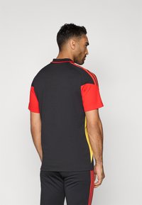 Camisa desportiva de manga curta com painéis em preto, vermelho e amarelo, colarinho clássico e tecido texturizado com realces contrastantes nos ombros.