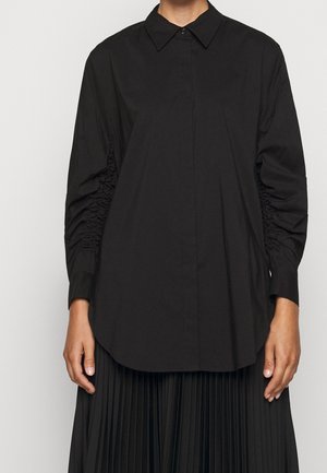 Camiseta negra de manga larga con detalles fruncidos en las mangas, cuello amplio y tapeta de botones oculta, combinada con una falda plisada.