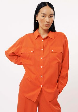 Chemise orange à manches longues avec boutonnage sur le devant, deux poches poitrine et une coupe décontractée, fabriquée à partir d'un tissu lisse avec un léger éclat.