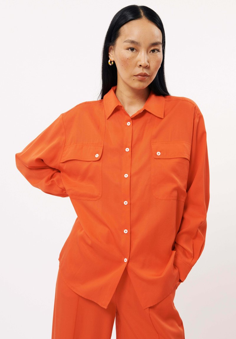 Chemise orange à manches longues avec boutonnage sur le devant, deux poches poitrine et une coupe décontractée, fabriquée à partir d'un tissu lisse avec un léger éclat.