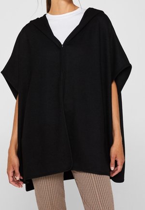 Woman wearing a loose black hooded poncho over a white top and checked slim-fit pants, standing with arms relaxed at sides.